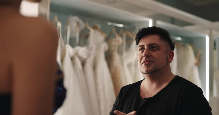 Clerk at dress shop talks to woman on elevated platform with wedding gowns hanging in rows in background