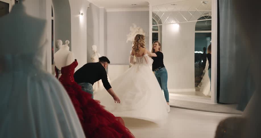 A man and a woman adjusting a wedding dress on a woman in a room with a wall and a mirror