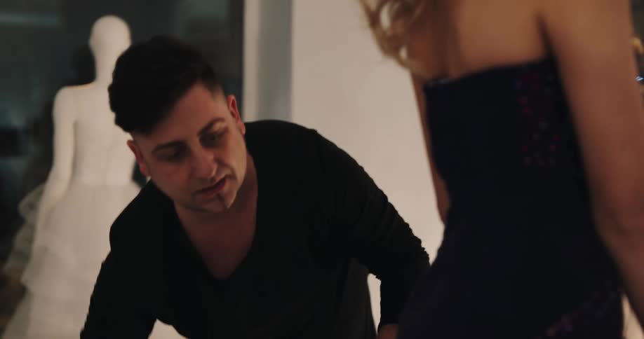 A stylist smooths out the train on an evening dress as a woman admires herself in the mirror