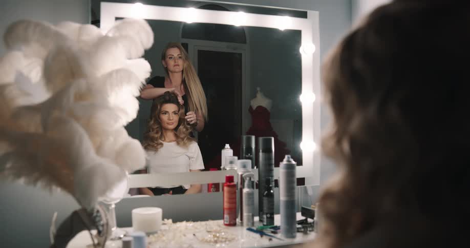 Blonder hairdresser puts on make up on woman with brown wavy hair in hairdresser salon