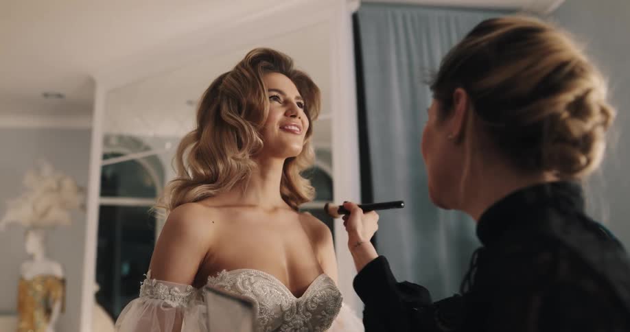 Makeup artist applies makeup to chest of woman in wedding gown as man straightens her train
