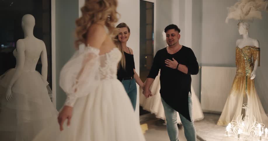 Woman with brown wavy hair shows wedding dress to man and woman holding hands in bridal shop