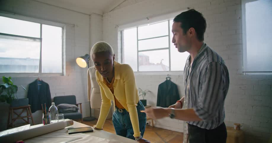 A man and woman discussing a design at a work table before walking over to a dress form