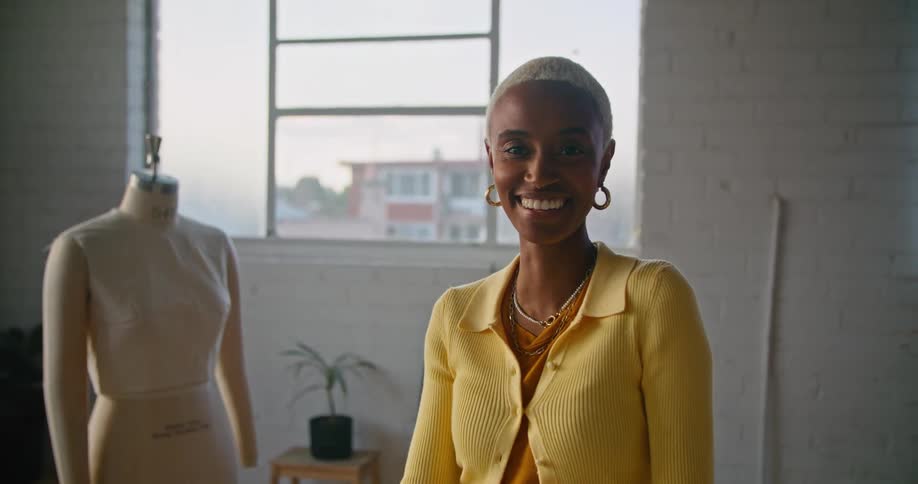 Female designer with short hair hoop earrings and yellow shirt smiles at camera in workshop
