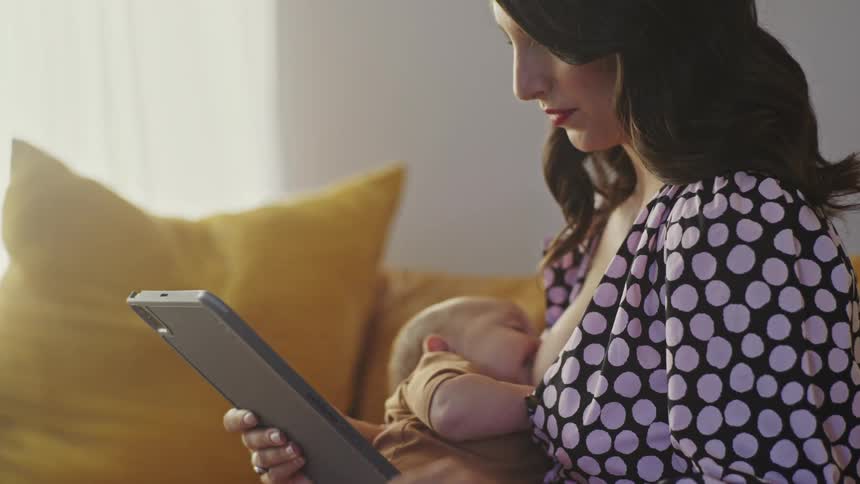 A woman scrolls through her tablet while breastfeeding her baby at home