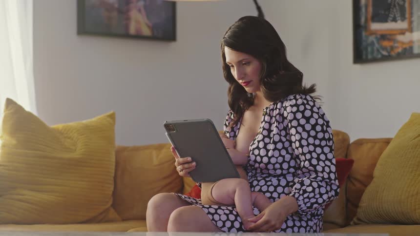 A woman breast feeds her baby while sitting on the sofa holding her tablet
