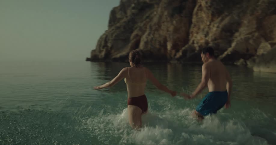 Man with blue swimming shorts and woman in red yellow bikini runs to the sea on pebble beach