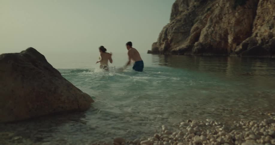 A man and a woman run across the beach into the water and splash each other