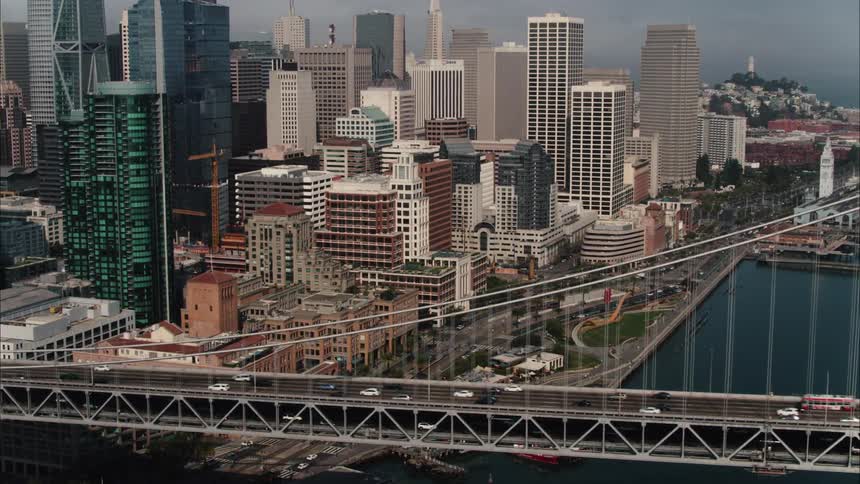 An aerial view of cars driving across a bridge in front of skyscrapers on the shore