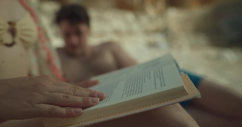 A woman sits on a beach paging through a book with a man laying beside her