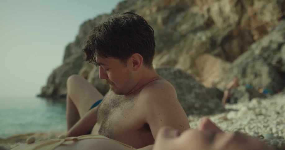 Woman laughs while lying on beach as smiling man places white pebbles on her belly