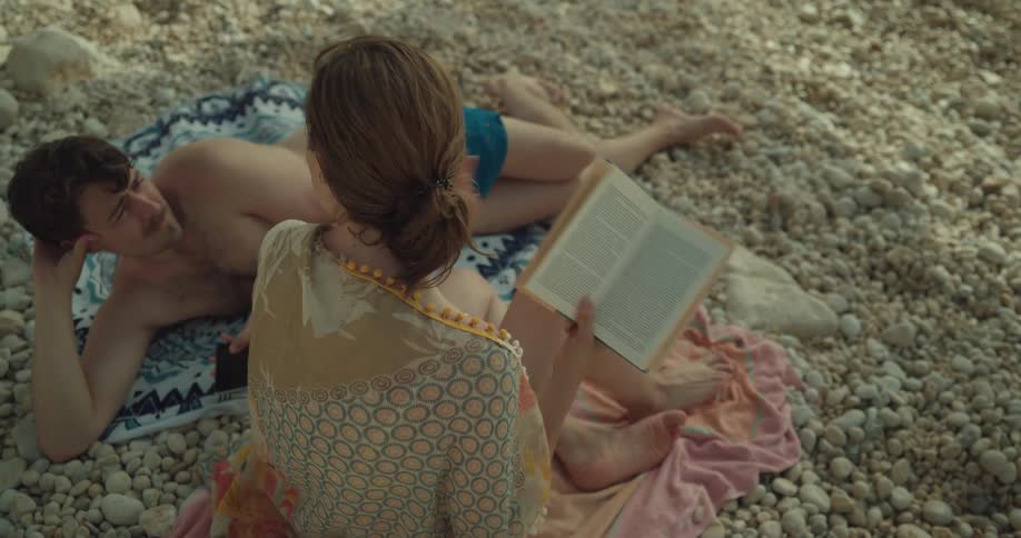 A woman sits on the beach reading a book and then leans forward and talk to a man who is lying beside her looking at her phone