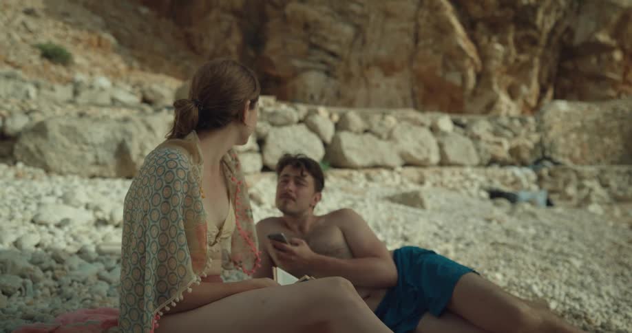 A man lies on the shore and uses his cell phone as he talks to a woman sitting and reading a book