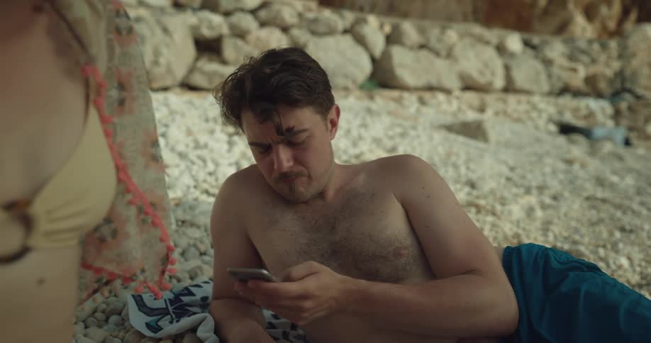 A man scrolls through his phone while his girlfriend reads a book on the beach