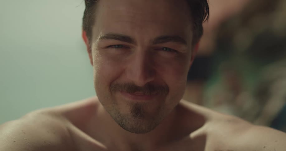 Close up of a happy young shirtless bearded man sitting on a beach looking at the camera