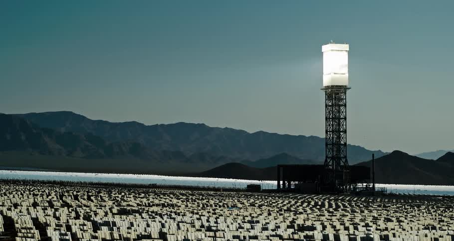 Light shines from box on top of tower bordering water and surrounded by small white panels with misty mountains in background
