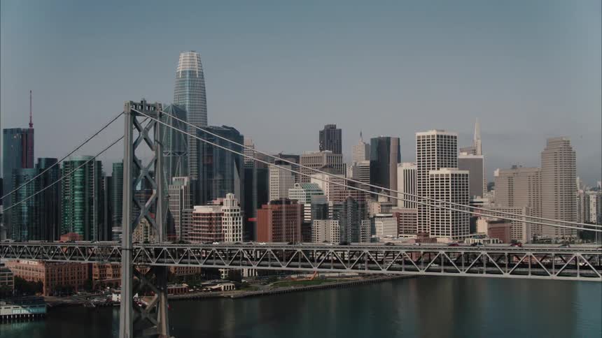 A city skyline with buildings and skyscrapers with a bridge in the foreground and the sky and clouds in the background