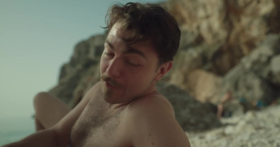 A man is lying on a beach wearing swimming trunks and smiling as he plays with the sand