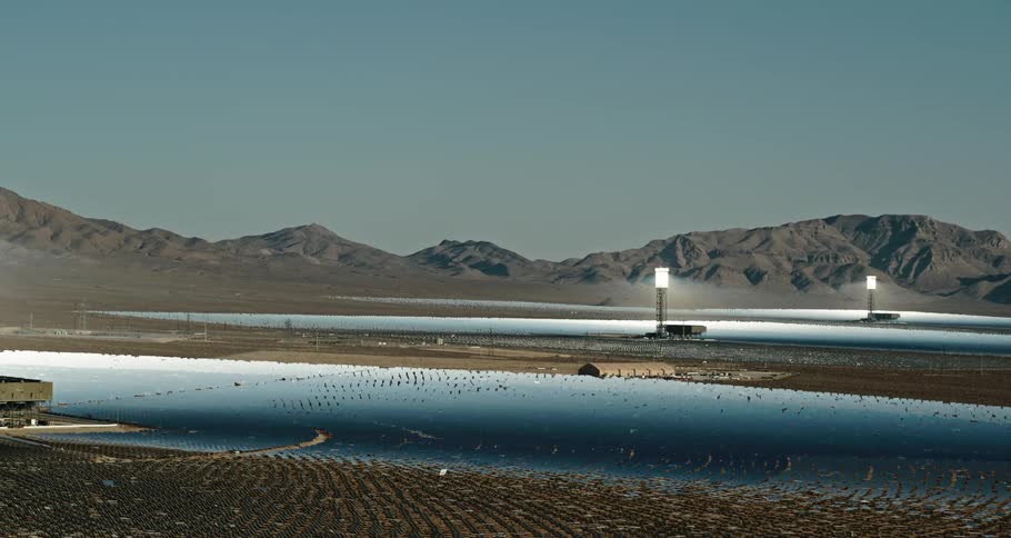 Series of bright lights sit on top of metal framed structures shining onto reflective blue surfaces in field stretching to mountains