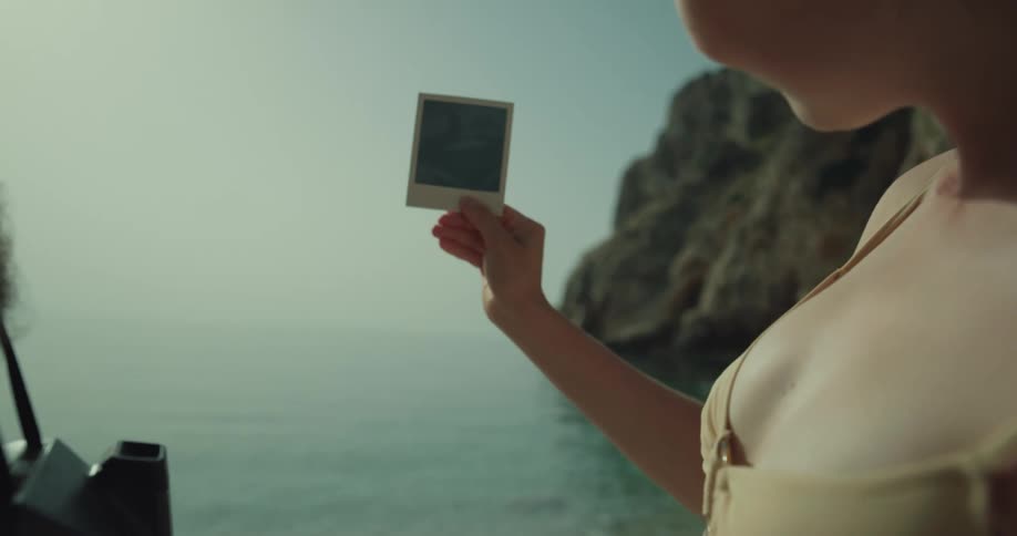 A woman shakes a polaroid picture by the water