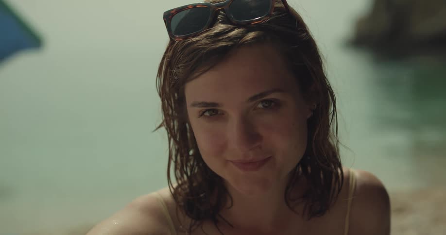 Woman puts sunglasses on head smiling while sitting on beach near water