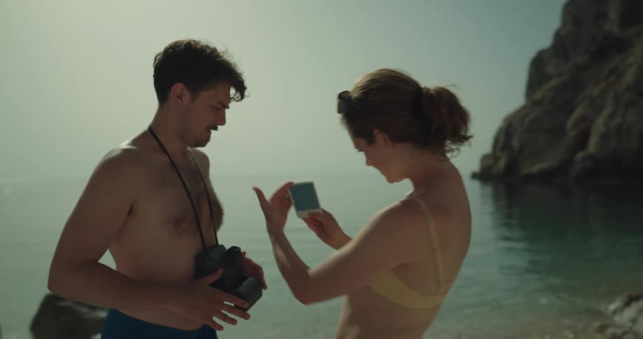 A man takes an instant style photograph and hands it to the woman he took the picture of while theyre standing near an ocean