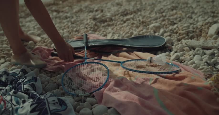 A woman picks up badminton rackets that are lying on a blanket on the beach