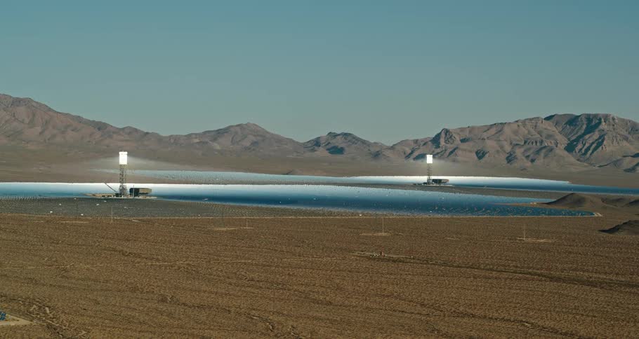 Two bright lights sitting on top of structure shine across metallic surface in plain with mountains in background under clear sky