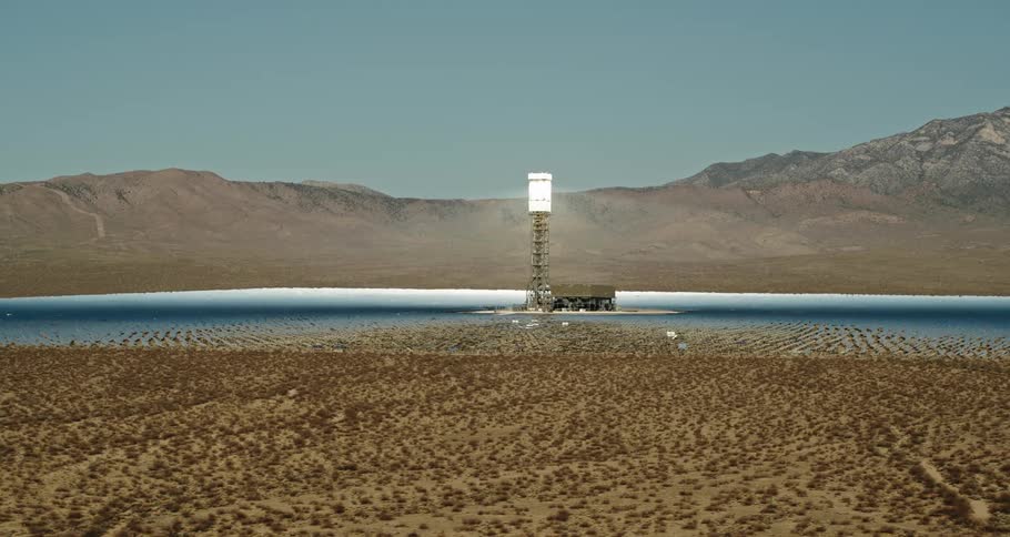 Light shines from top of tower set in metallic blue area between scrubland and mountain under blue sky