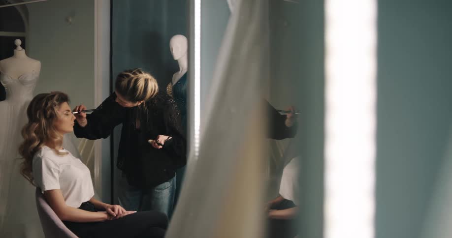 A woman sitting in a room as another woman applies makeup to her face with a mirror and a wall in the background