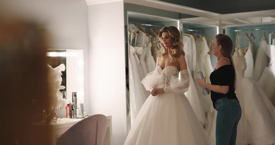 A woman stands in a wedding gown in a wedding studio with another woman fixing her hair
