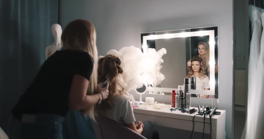 A woman in a room sitting on a chair as another woman does her hair with a wall and a mirror in the background