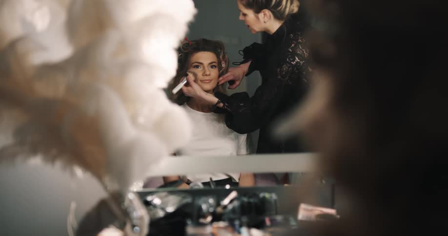 Woman with black shirt puts makeup on bride with brown wavy hair looking in mirror