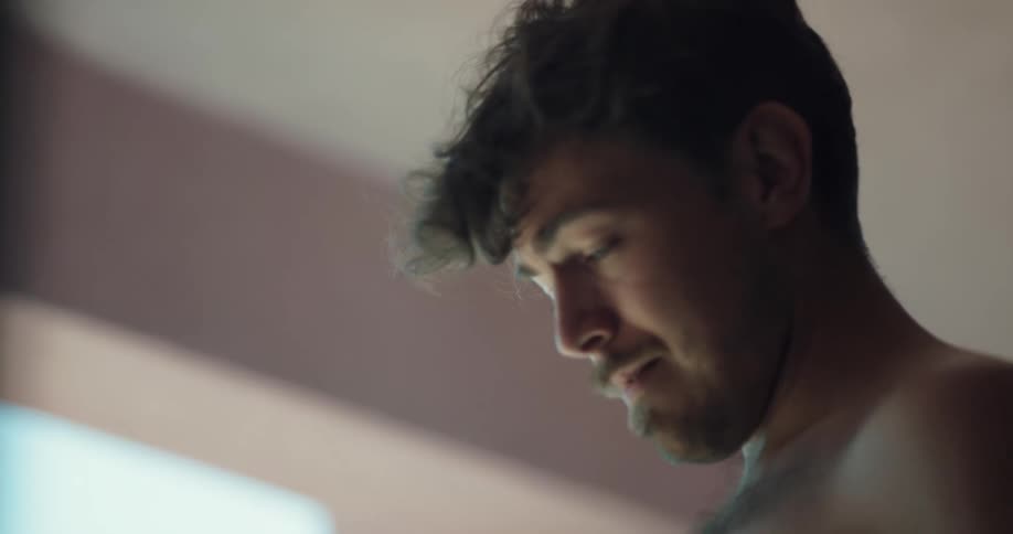 A bare chested man is looking around and rubs his hand across his forehead