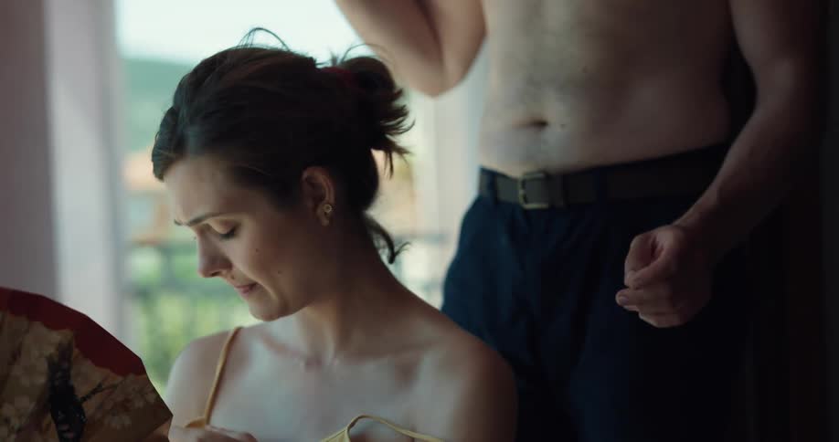 A man and a woman in a room with the woman fanning herself with a fan and a man applying a spray on her back with a wall and window in the background