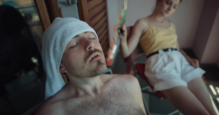 Woman uses fan to fan herself and bare chested man wearing towel on his head