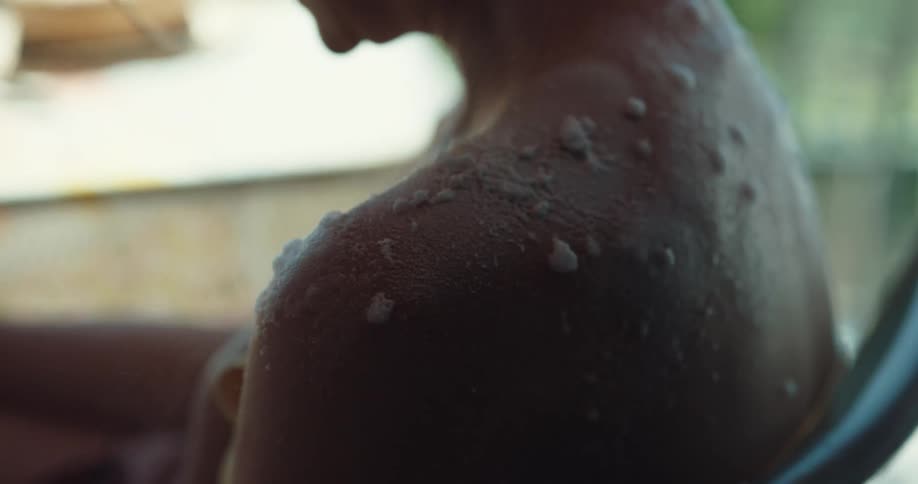 A woman sitting on a chair at a table as another person sprays a white cream on her skin