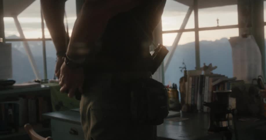 Man with cap black shirt and brown pants puts on walky talky in room overlooking mountains