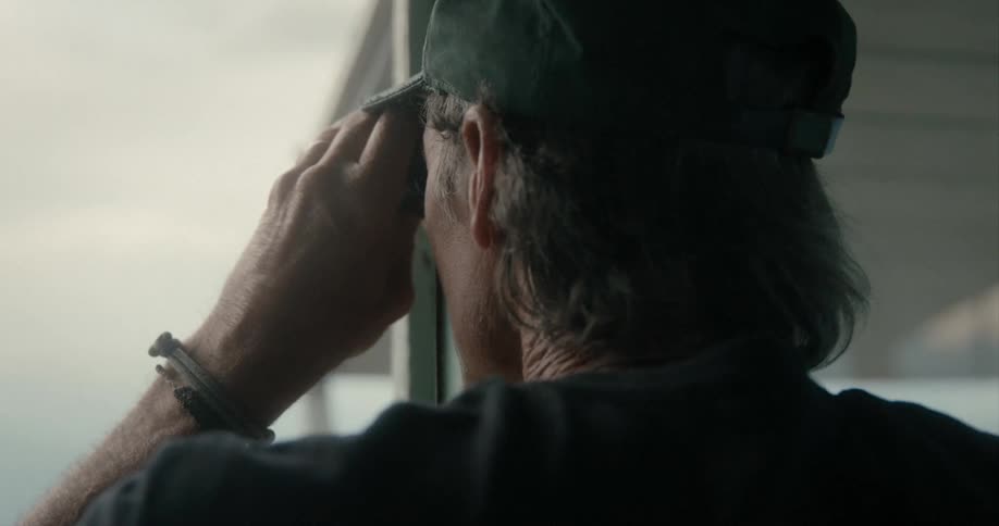 A man with a hat looking through binoculars with the sky and clouds in the background