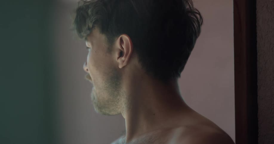 A bare chested man puts his hand on his forehead and looks around
