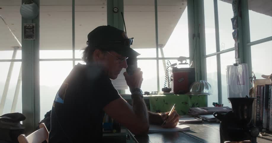 A man sits at a desk as he talks on a walkie and writes on a paper