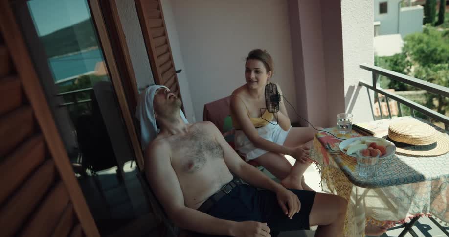 Shirtless man puts wet towel on his head while woman uses hand fan on the balcony getting exhausted from heat