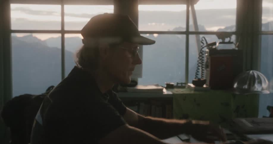 Man with cap black polo shirt sits down at table wearing glasses to read notebook in room overlooking distant mountains
