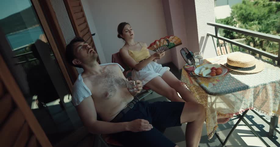 Couple sits on shady balcony fanning themselves and drinking water
