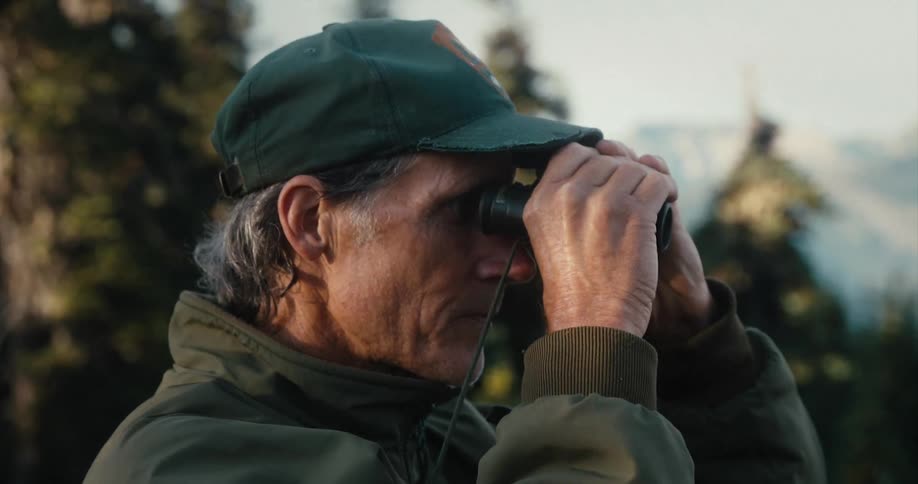Man wearing baseball cap standing on a forest in the mountains looking through binoculars