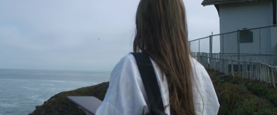Young woman holding a tablet stands near a fence in front of the ocean