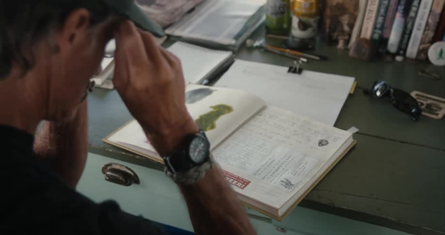 Man wearing baseball cap puts on glasses to read a book over a desk