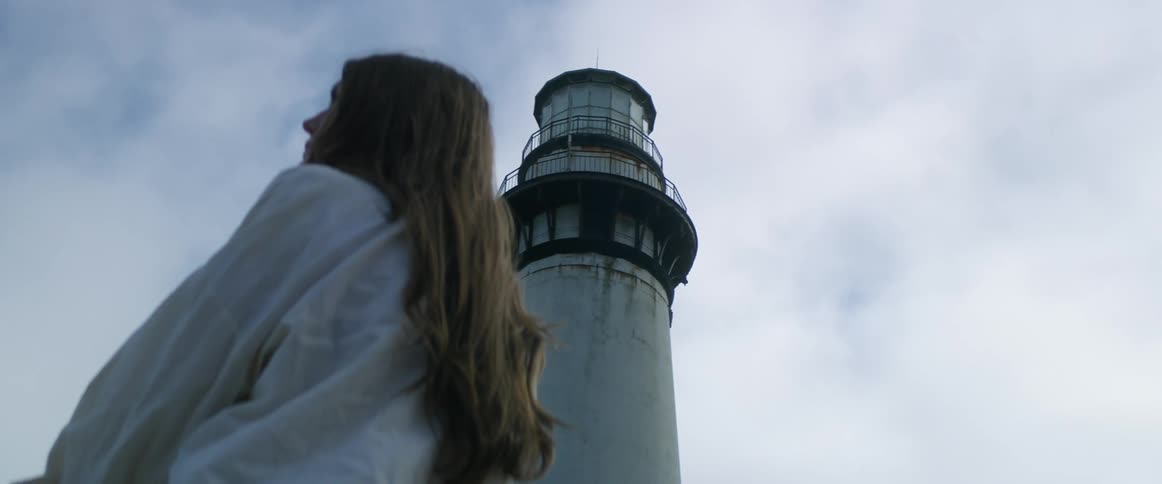Woman wearing lose flowing blouse stands near lighthouse under heavily cloudy sky
