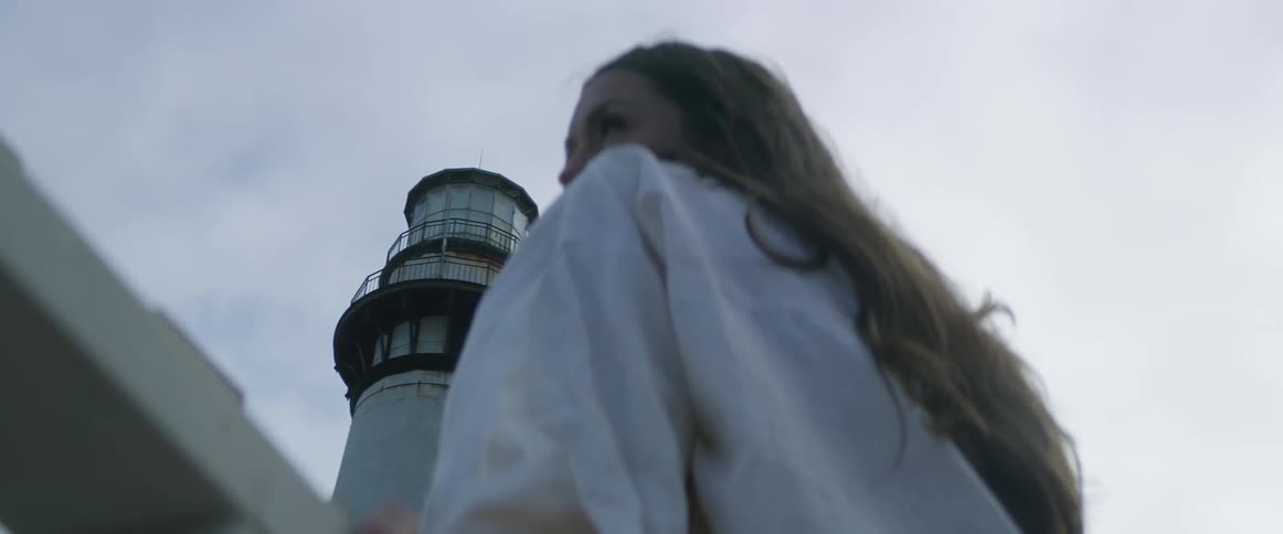 A woman with long brown hair is leaning on a railing and a lighthouse is in the distance
