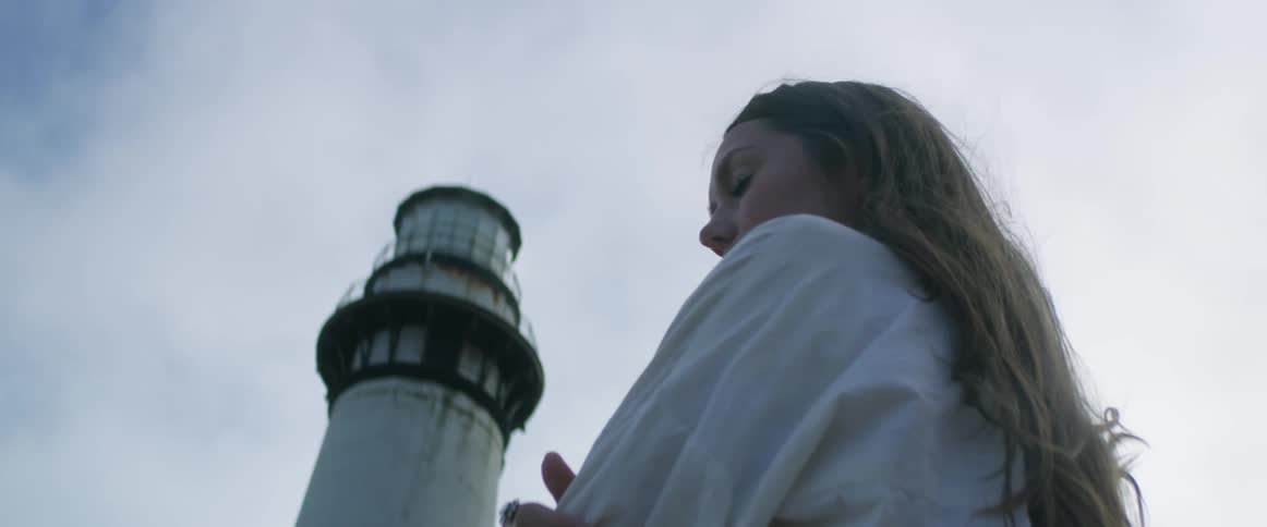 A woman stands with her arms crossed in front of a lighthouse